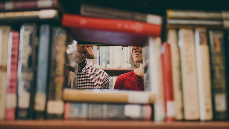 Foreground: books on a shelf stacked to form a square hole; Background, through the hole: a man and a woman smiling at each other, but you can only see their smiles and chins and their shoulders.