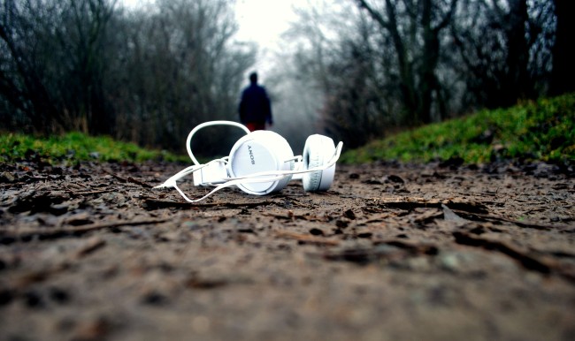 Photo from the ground, focused on foregrounded pair of white over-ear headphones lying on the ground, on a dirt path. Path is lined with grass and many dead trees. A human, wearing a black hoodie, is walking away down the middle of the path.